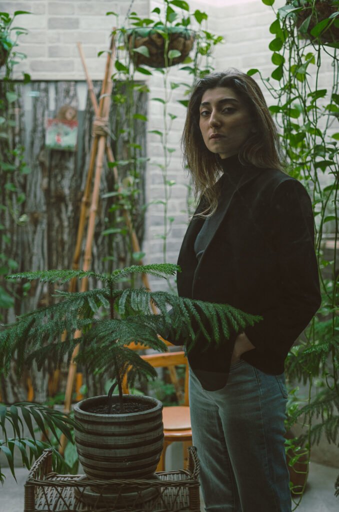 Mubie standing behind a potted evergreen in a plant-filled interior