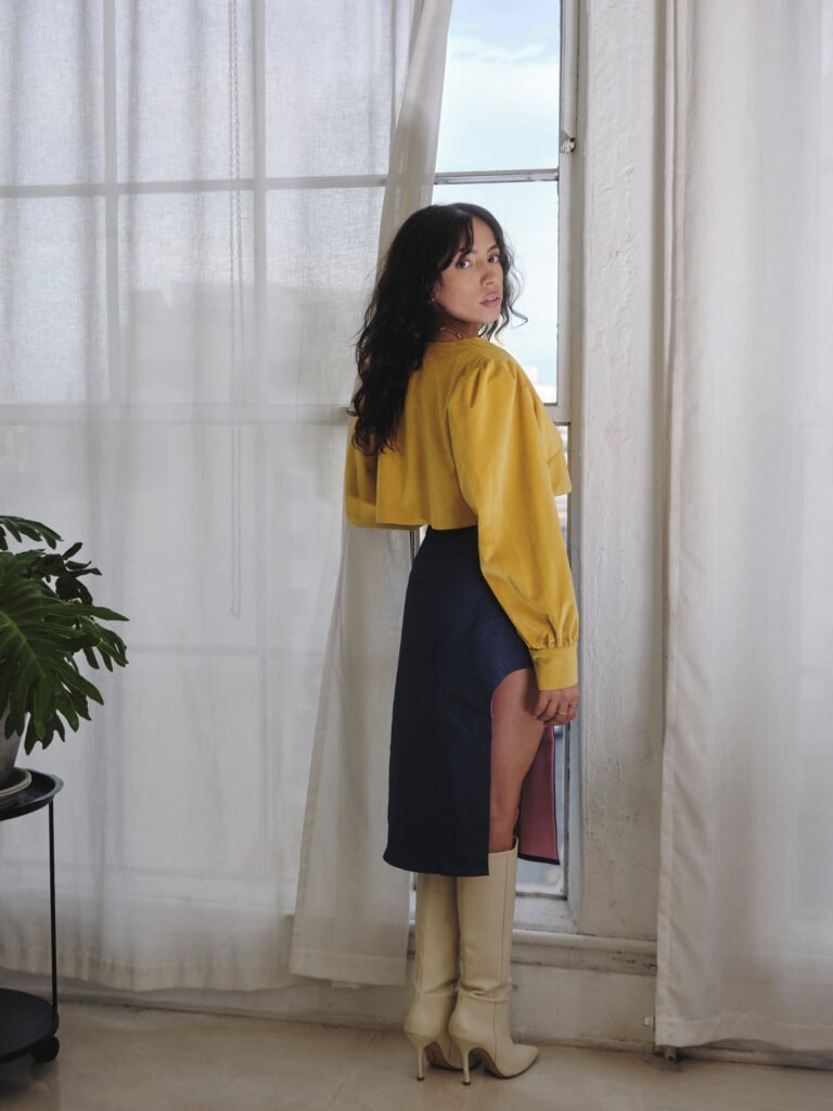 Neena Roe standing by a bright window in a yellow top and blue skirt