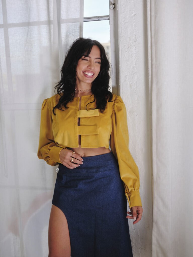 Neena Roe smiling beside a bright window in a yellow top and blue skirt