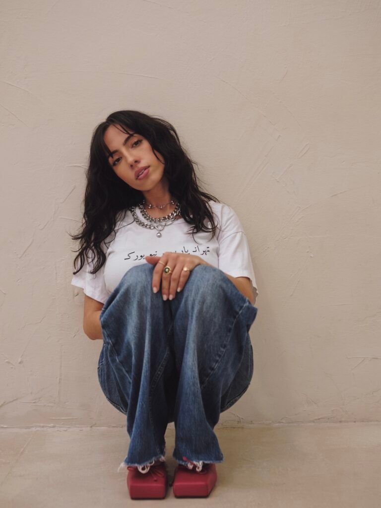 Neena Roe seated against a textured cream wall wearing a white T-shirt and jeans