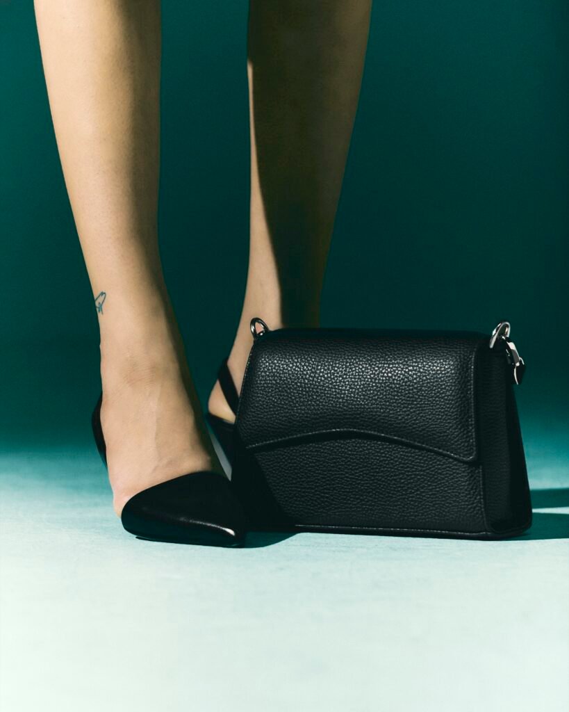 Black leather work bag beside a model's shoes against a teal studio backdrop