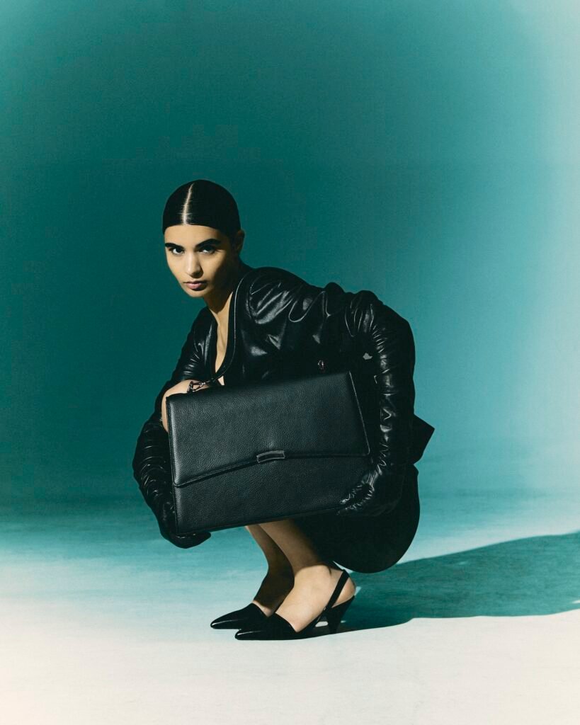 Model crouching while holding a large black leather work bag