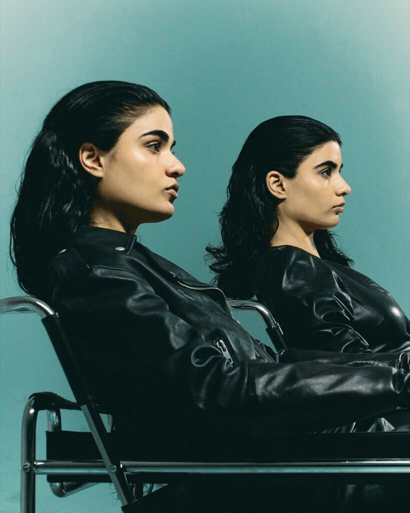 Two models in black leather outfits seated against a teal studio backdrop