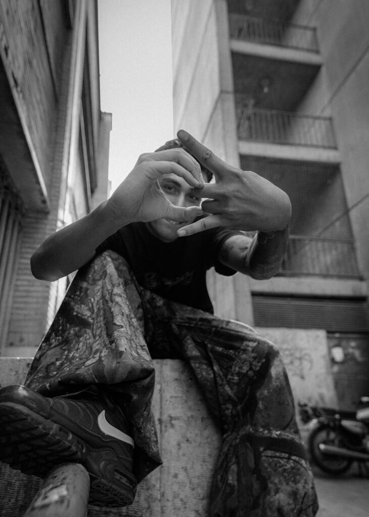 A black and white portrait of Emad in a back street in Tehran making pop-culture hand gestures at he camera